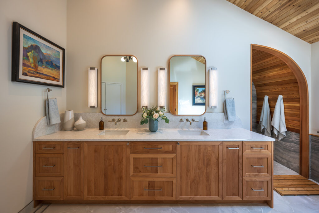 Large, double vanity with quartz counters and alabaster sconces, blue vase with flowers, wall mounted faucets, in satin nickel finish, custom wood framed mirrors with rounded edges.