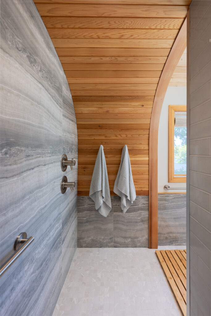 Curbless shower with curved, wood paneled ceiling, satin nickel accents, porcelain tile floors and walls, grab bar and towel hooks.